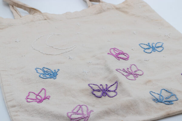 Naturfarbener handbestickter Beutel mit zarten Schmetterlingen im Fineline-Stil am unteren Bereich. Die Schmetterlinge sind in pastellfarbenem Garn in Rosa, Blau und Flieder gestickt. Darüber befinden sich ein Halbmond und funkelnde Sterne in weißem Garn, die dem Design eine träumerische und himmlische Note verleihen.