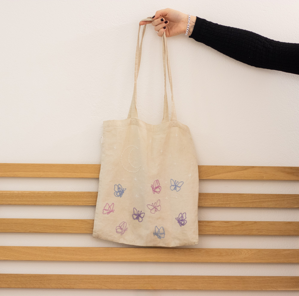 Naturfarbener handbestickter Beutel mit zarten Schmetterlingen im Fineline-Stil am unteren Bereich. Die Schmetterlinge sind in pastellfarbenem Garn in Rosa, Blau und Flieder gestickt. Darüber befinden sich ein Halbmond und funkelnde Sterne in weißem Garn, die dem Design eine träumerische und himmlische Note verleihen.