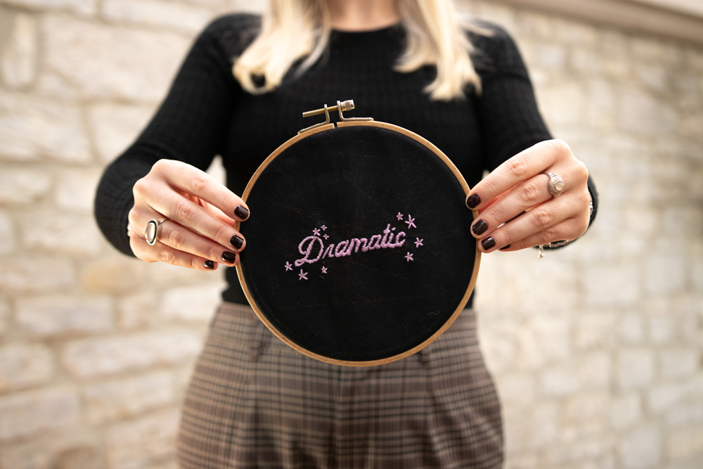 Rundes Stickbild in einem Stickrahmen mit schwarzem Stoff als Hintergrund. In fliederfarbenem Garn ist das Wort „Dramatic“ in eleganter Schnörkelschrift gestickt. Um den Schriftzug herum sind kleine Sterne verteilt, die das Design dezent schmücken und eine verträumte Stimmung erzeugen.