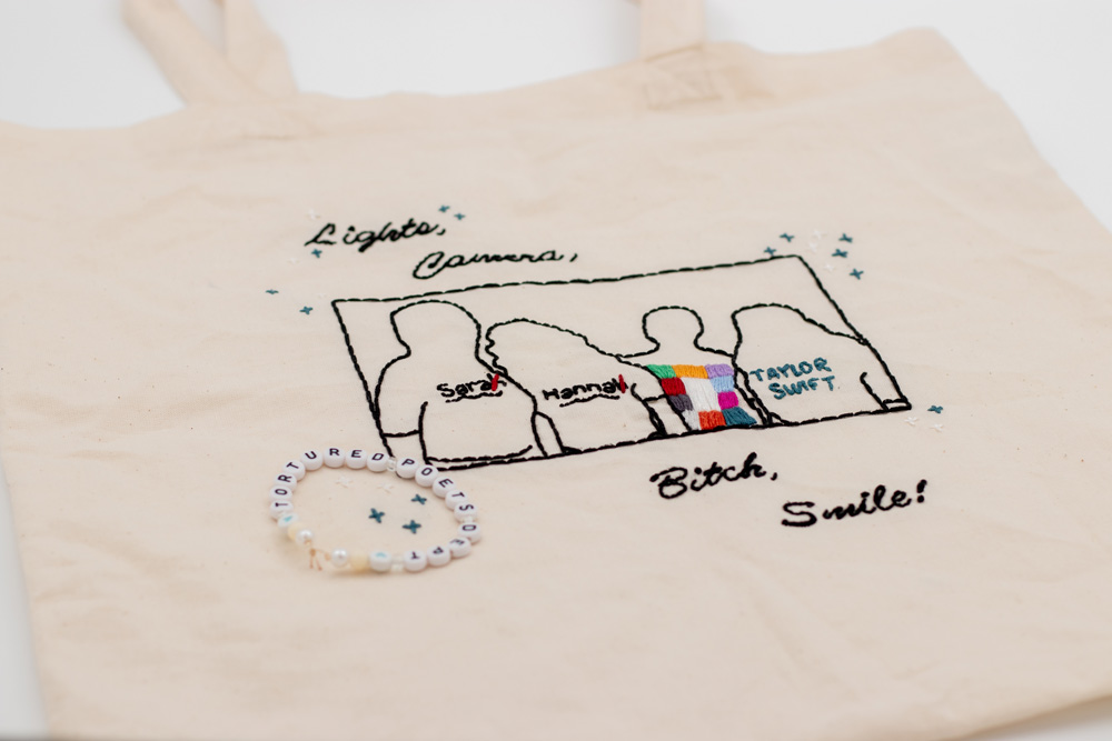Naturfarbener handbestickter Beutel mit einem Fineline-Motiv, das vier Freundinnen in ihren individuellen Eras Tour Outfits zeigt – inspiriert von einem Foto der Tour. Das Motiv wird von einem zarten Rahmen und kleinen gestickten Sternen umgeben. Ober- und unterhalb des Designs befindet sich in eleganter Schnörkelschrift die Liedzeile „Lights, Camera, Bitch, Smile“, die das lebhafte und besondere Flair der Eras Tour einfängt.