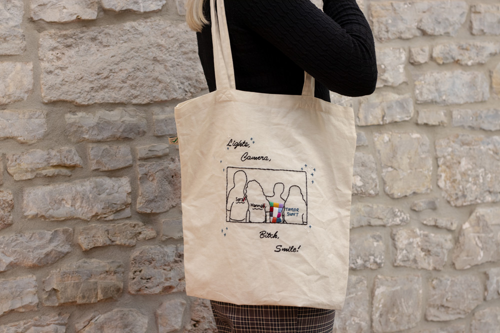 Naturfarbener handbestickter Beutel mit einem Fineline-Motiv, das vier Freundinnen in ihren individuellen Eras Tour Outfits zeigt – inspiriert von einem Foto der Tour. Das Motiv wird von einem zarten Rahmen und kleinen gestickten Sternen umgeben. Ober- und unterhalb des Designs befindet sich in eleganter Schnörkelschrift die Liedzeile „Lights, Camera, Bitch, Smile“, die das lebhafte und besondere Flair der Eras Tour einfängt.