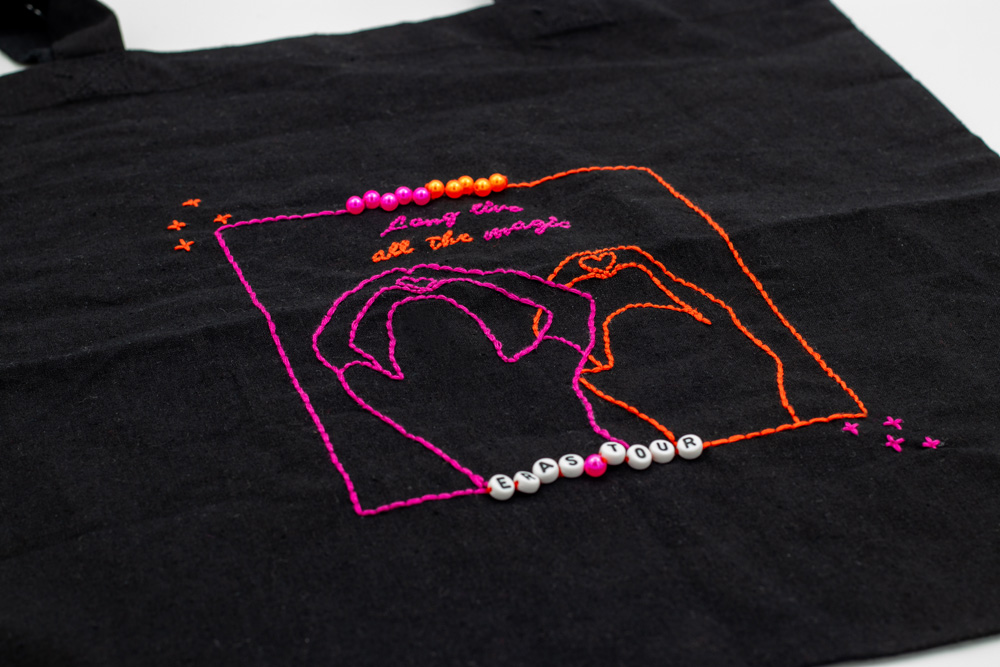 Schwarzer handbestickter Beutel mit einem Fineline-Motiv in Pink und Orange, das zwei Freundinnen von hinten zeigt, die mit ihren Händen ein Herz formen – inspiriert von einem Moment auf der Eras Tour. Oberhalb der Hände ist in eleganter Schnörkelschrift die Liedzeile "Long live all the magic" gestickt. Das Motiv ist mit einem filigranen Rahmen in denselben Farben umgeben, der mit kleinen Perlen verziert ist. Am unteren Rand des Rahmens bilden die Perlen im Stil eines Freundschaftsarmbands das Wort „Eras Tour“. Basierend auf der Vorlage eines Fotos von der Eras Tour.