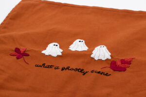 Rostbrauner handbestickter Kissenbezug mit drei kleinen weißen Geistern, die schwarze Augen haben und nebeneinander angeordnet sind. Links und rechts der Geister befindet sich je ein großes, weinrotes Blatt. Unterhalb des Motivs ist in eleganter Schnörkelschrift die Liedzeile „what a ghostly scene“ gestickt, was dem Design eine herbstliche und mystische Atmosphäre verleiht.