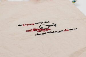 Naturfarbener handbestickter Beutel mit einem umgekippten Weinglas in schwarzem Garn, aus dem burgunderrote Weinpfützen im Fineline-Stil fließen. Das Motiv wird von der Liedzeile „The burgundy on my t-shirt when you splashed your wine onto me“ umrahmt, die in einer eleganten Schrift gestickt ist. Ein detailreiches Design, das Musik und Kunst verbindet.