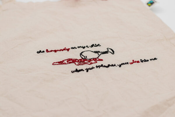 Naturfarbener handbestickter Beutel mit einem umgekippten Weinglas in schwarzem Garn, aus dem burgunderrote Weinpfützen im Fineline-Stil fließen. Das Motiv wird von der Liedzeile „The burgundy on my t-shirt when you splashed your wine onto me“ umrahmt, die in einer eleganten Schrift gestickt ist. Ein detailreiches Design, das Musik und Kunst verbindet.