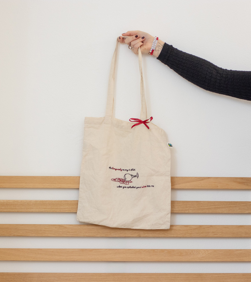 Naturfarbener handbestickter Beutel mit einem umgekippten Weinglas in schwarzem Garn, aus dem burgunderrote Weinpfützen im Fineline-Stil fließen. Das Motiv wird von der Liedzeile „The burgundy on my t-shirt when you splashed your wine onto me“ umrahmt, die in einer eleganten Schrift gestickt ist. Ein detailreiches Design, das Musik und Kunst verbindet.