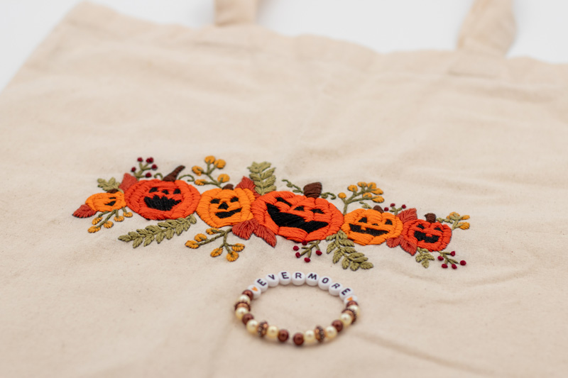 Naturfarbener handbestickter Beutel mit fünf Kürbissen in verschiedenen Orangetönen, die geschnitzte Gesichter tragen. Die Kürbisse sind von zarten grünen Pflanzenranken umgeben und verleihen dem Design einen herbstlichen und verspielten Charme. Inspiriert von Taylor Swifts Album "evermore".