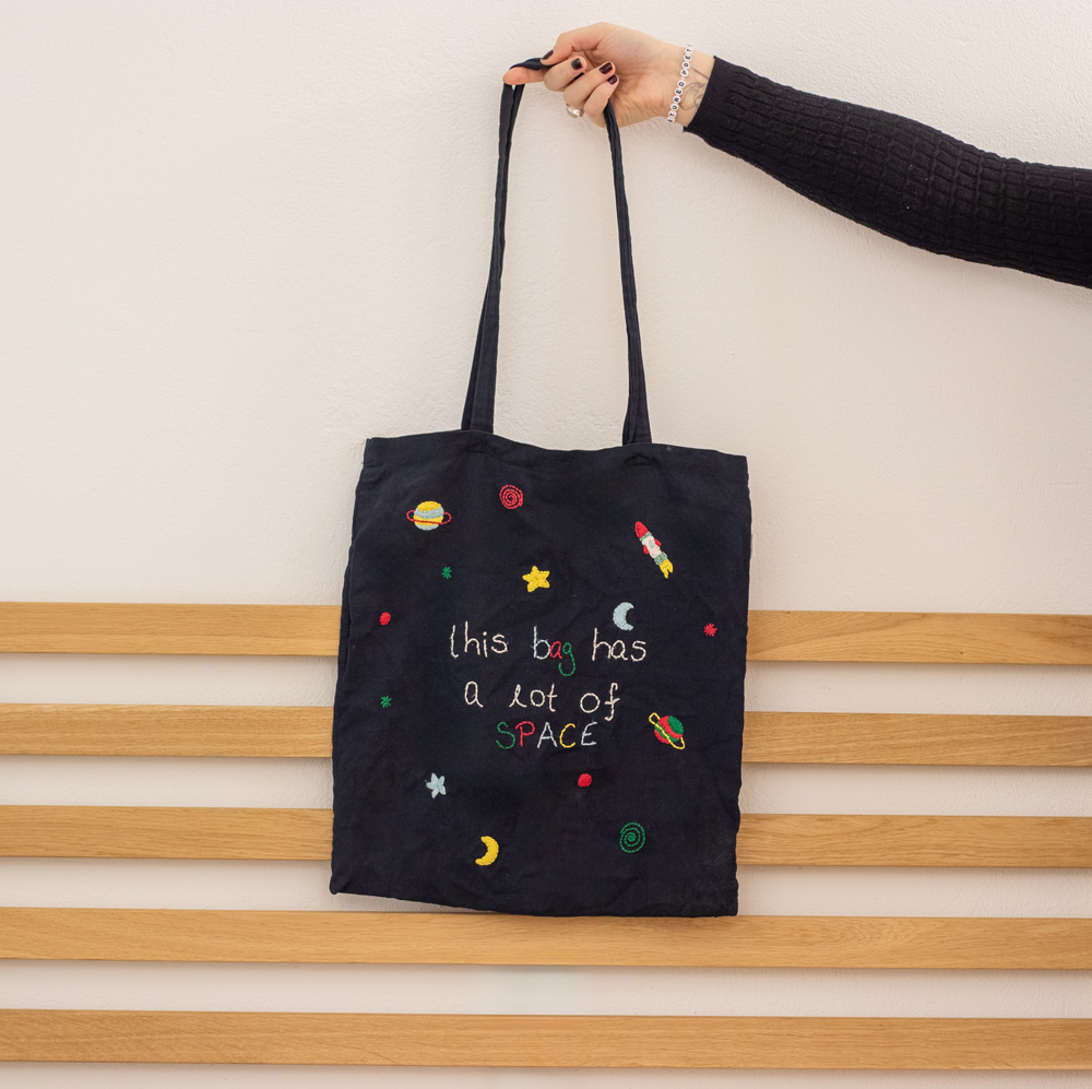 Schwarzer handbestickter Beutel mit verschiedenen kleinen Weltraum-Motiven, darunter eine Rakete, Planeten, Sterne und Monde, die locker über den Beutel verteilt sind. In der Mitte befindet sich der humorvolle Schriftzug „this bag has a lot of SPACE“, der das Thema des Designs aufgreift. Ein kreatives und witziges Accessoire für Weltraumfans.