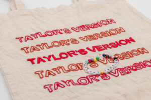 Naturfarbener handbestickter Beutel mit dem Schriftzug „Taylor's Version“ fünf Mal untereinander in Großbuchstaben gestickt. Die äußeren Zeilen sind in weinrot gehalten, während die mittlere Zeile in rostrot hervorsticht und dem Design eine besondere Dynamik verleiht. Ein minimalistisches, aber ausdrucksstarkes Motiv für Fans.