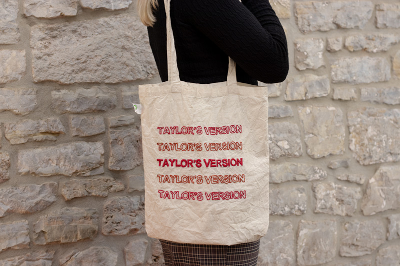 Naturfarbener handbestickter Beutel mit dem Schriftzug „Taylor's Version“ fünf Mal untereinander in Großbuchstaben gestickt. Die äußeren Zeilen sind in weinrot gehalten, während die mittlere Zeile in rostrot hervorsticht und dem Design eine besondere Dynamik verleiht. Ein minimalistisches, aber ausdrucksstarkes Motiv für Fans.